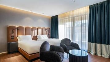 A modern hotel room with a large bed, two armchairs, a small round table, and a nightstand with a lamp.