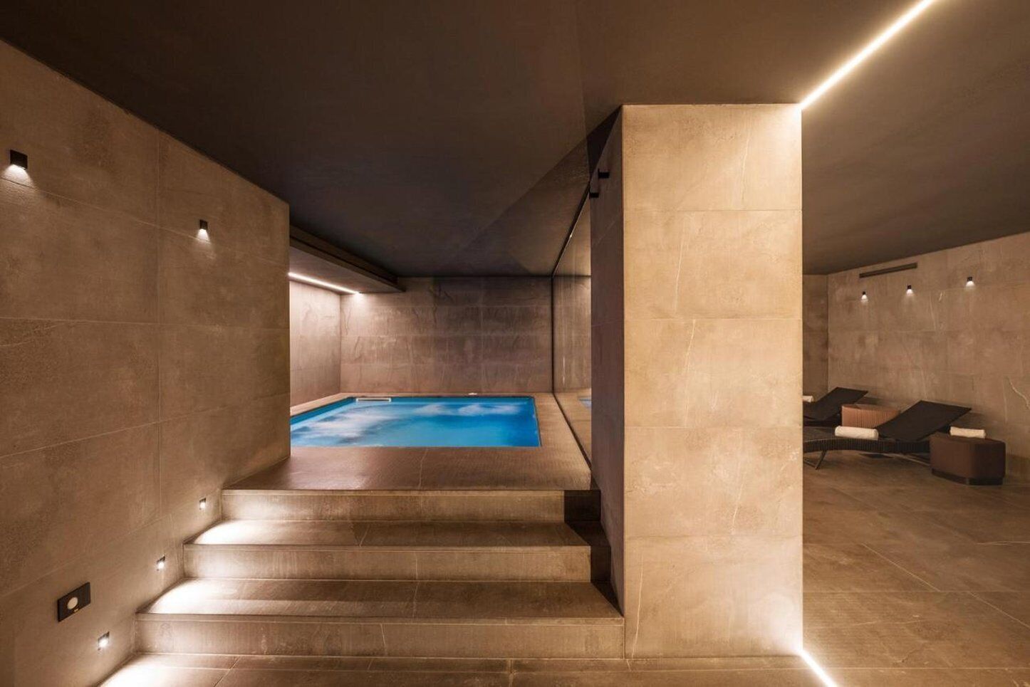 Una piscina interna moderna con superficie d'acqua blu trasparente, circondata da un design minimalista con pareti in pietra e una scala che conduce a una zona relax con lettini.