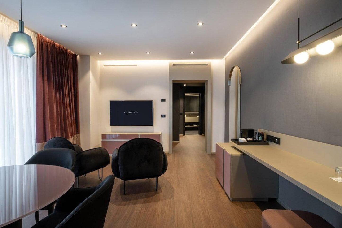 A modern hotel room with a flat-screen TV, a dining area with a round table and chairs, a kitchenette, and a hallway leading to a bathroom.