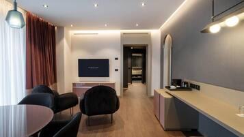 A modern hotel room with a flat-screen TV, a dining area with a round table and chairs, a kitchenette, and a hallway leading to a bathroom.