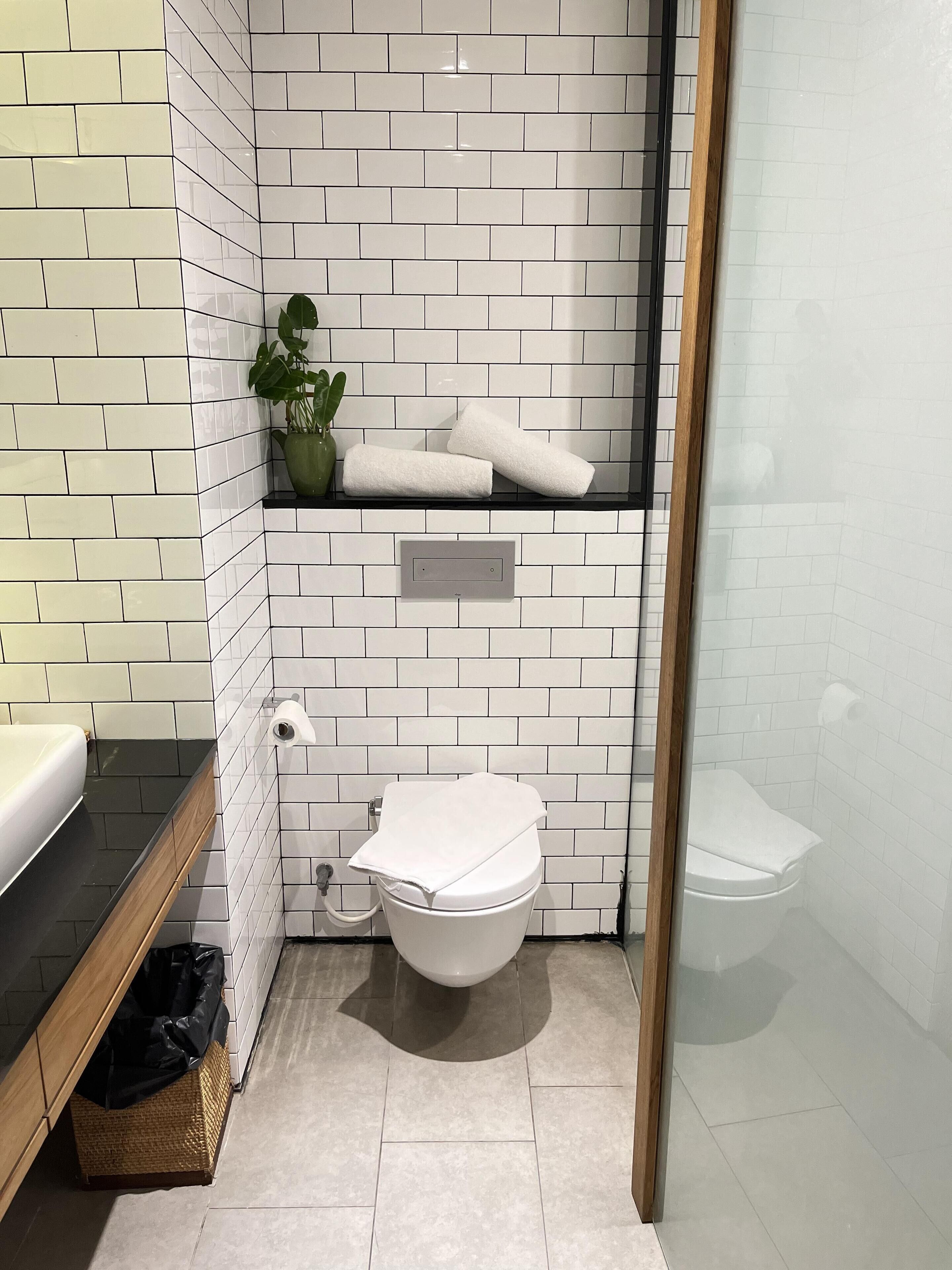 Egy modern fürőszoba fehér, téglalap alakú csempékkel, fekete mosdólappal, fehér WC-vel, és egy kis polccal, amelyen törölközők és egy virágos növény található.