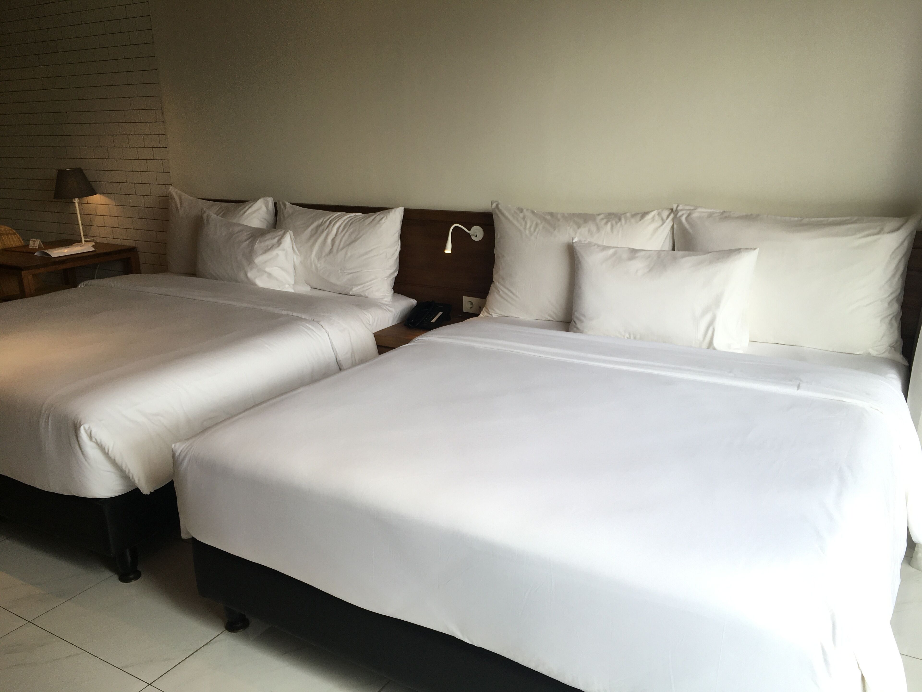 Habitación de hotel con dos camas individuales, una mesita de noche con lámpara y una pared de ladrillo.