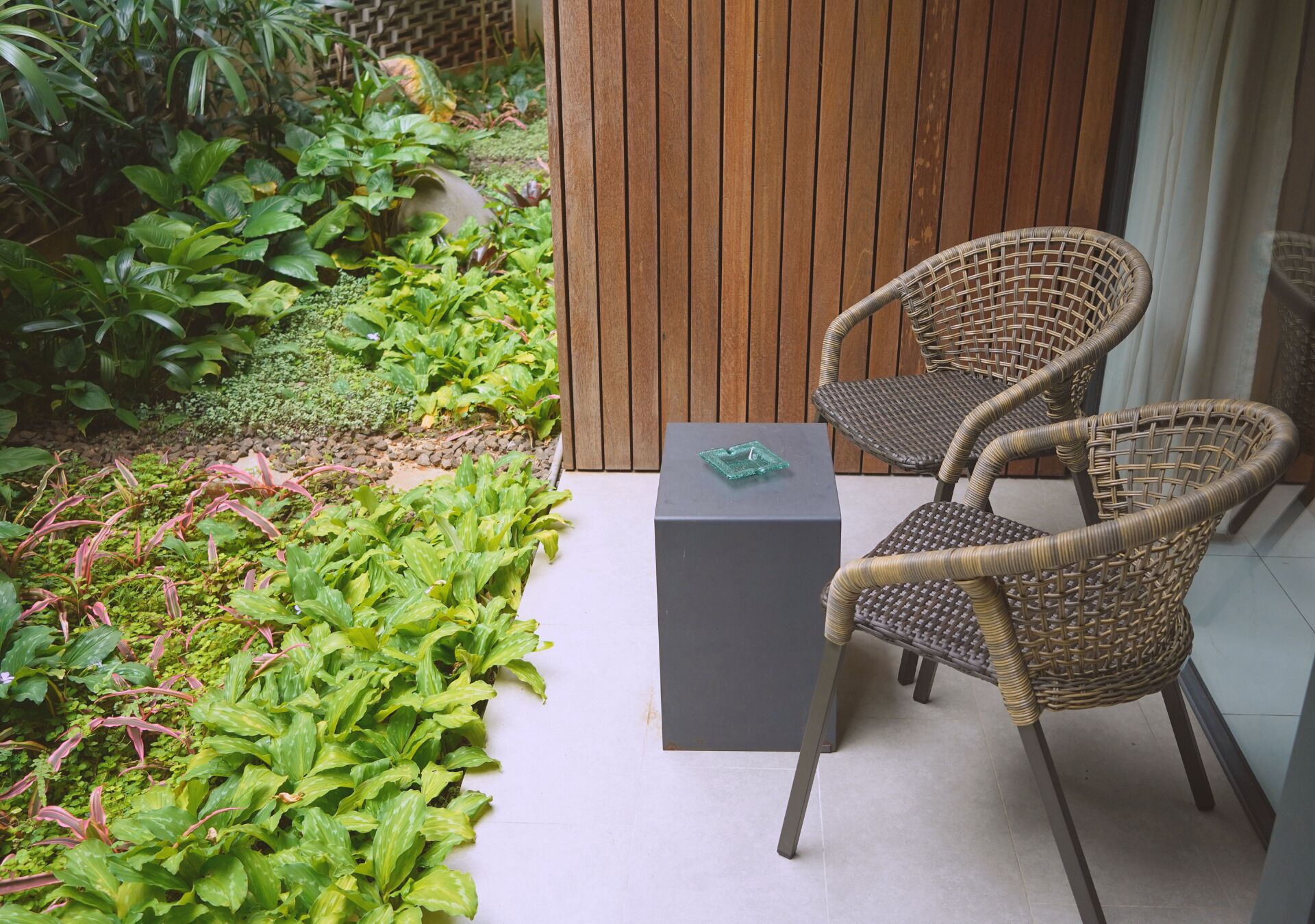 A small outdoor seating area with wicker chairs and a table, surrounded by lush greenery and a wooden fence.