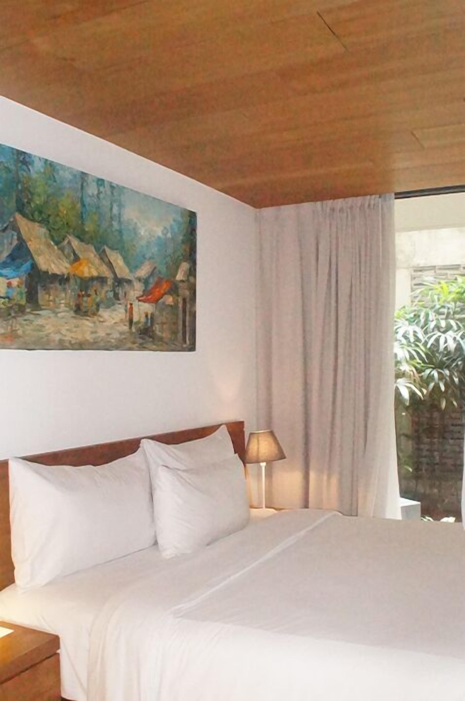 A hotel room with a bed, a painting of a village scene, a lamp, and a window with curtains.