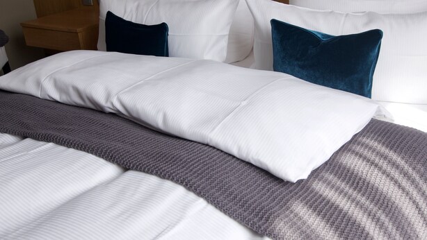 A neatly made bed with white and gray bedding, a wooden headboard, and a bedside table with a lamp. - Hotel Tari (Hoechberg)