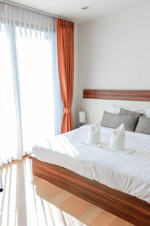 A hotel room with a large bed, wooden headboard, white bedding, and a view of the outdoors through sheer curtains.