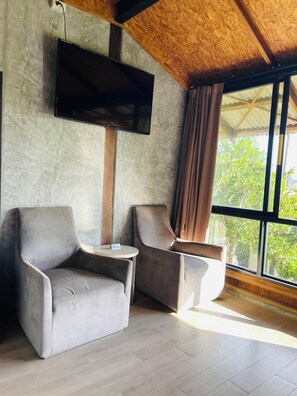 A room with two armchairs, a small table, a flat-screen TV mounted on the wall, and a large window offering a view of greenery outside.