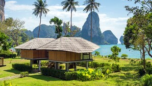 Une maison en bois avec un toit de chaume, entourée d’une végétation luxuriante et de palmiers, qui offre une vue sur une mer calme et des montagnes lointaines.