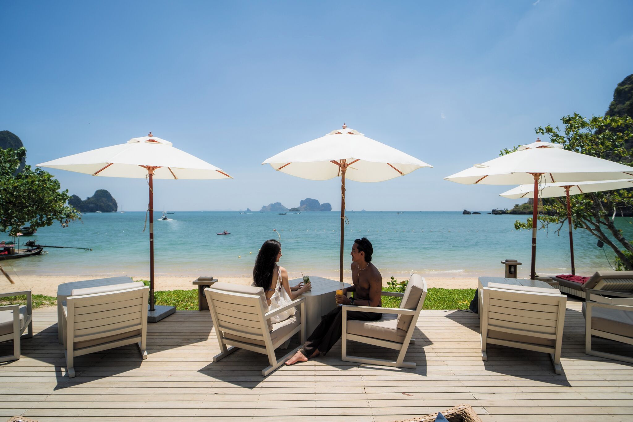 Une terrasse avec des sièges et des parasols, donnant sur une plage aux eaux cristallines et offrant une vue sur des îles lointaines.