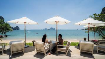 Une terrasse avec des sièges et des parasols, donnant sur une plage aux eaux cristallines et offrant une vue sur des îles lointaines.
