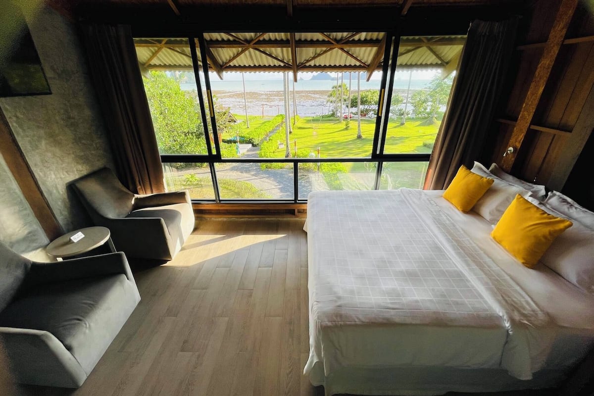 Un dormitorio con una cama grande, dos sillones y vistas a un paisaje verde a través de amplios ventanales.