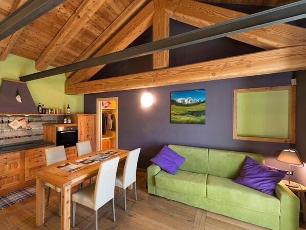 A cozy room with a wooden ceiling, a green sofa, a dining table with chairs, and a kitchen area with cabinets and a stove. - Agriturismo Gli Elfi (Saint-Christophe)