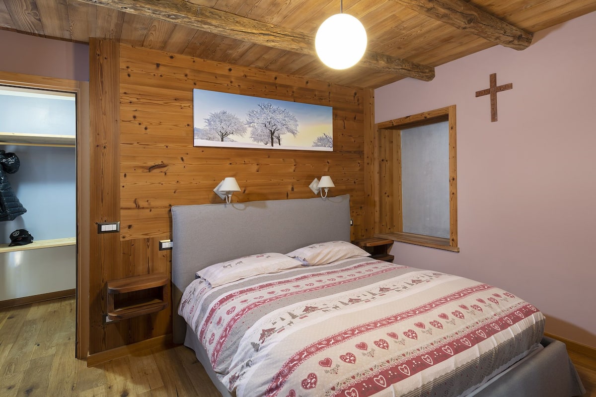 Ein Schlafzimmer mit Holzboden, einem Bett mit gemusterten Bettbezügen, zwei Nachttischlampen, einem Baumgemälde, einem Kreuz an der Wand und einem Fenster mit Jalousie.
