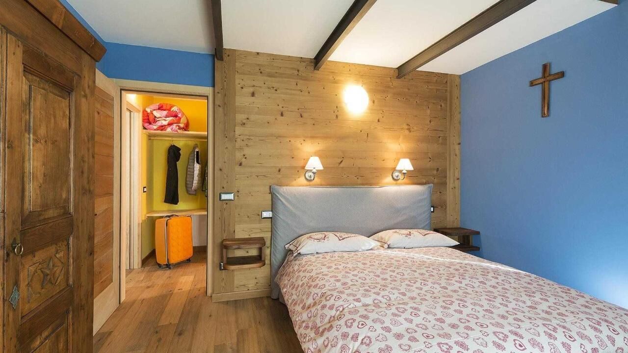 A bedroom with a wooden headboard, a bed with patterned bedding, a nightstand with two wall-mounted lamps, a wooden wardrobe, and a cross on the wall.