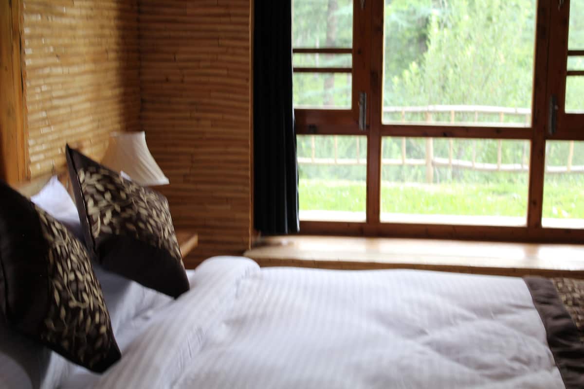 Una cama con ropa de cama blanca y almohadas estampadas, un cabecero de madera, una ventana con vistas a la vegetación y una mesita de noche con una lámpara.
