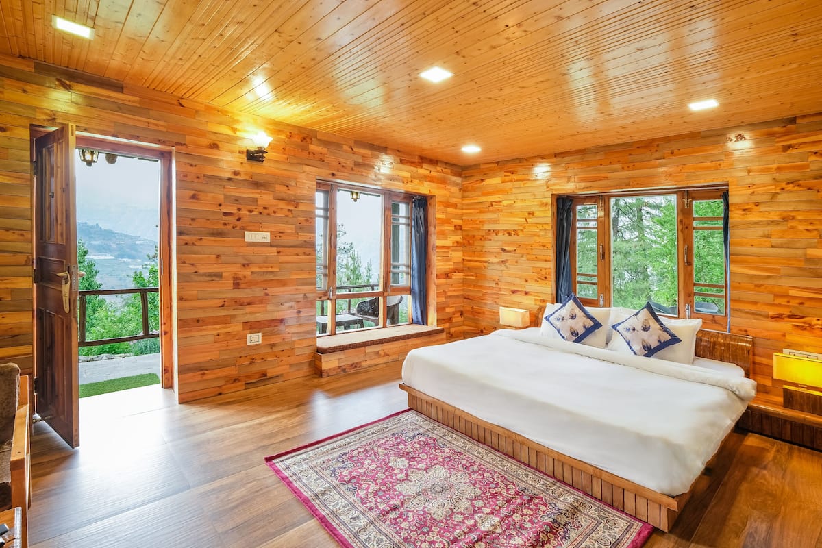 Un dormitorio de cabaña de madera con una cama grande, dos ventanas y vistas al exterior.