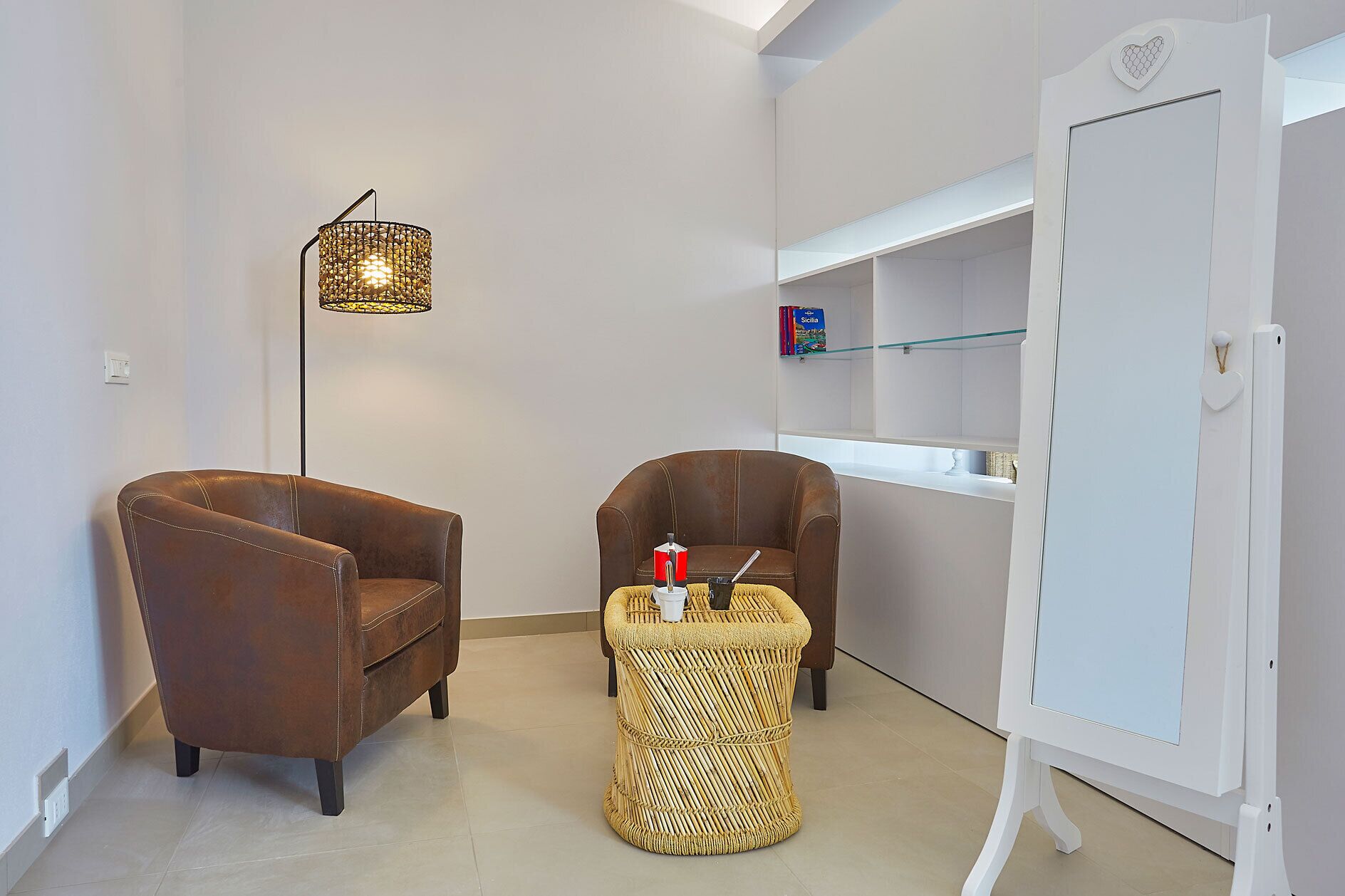 Quarto com duas poltronas de couro, um banquinho de vime, uma luminária de chão, uma lousa e uma prateleira com uma caixa identificada como 'Sicilia'.
