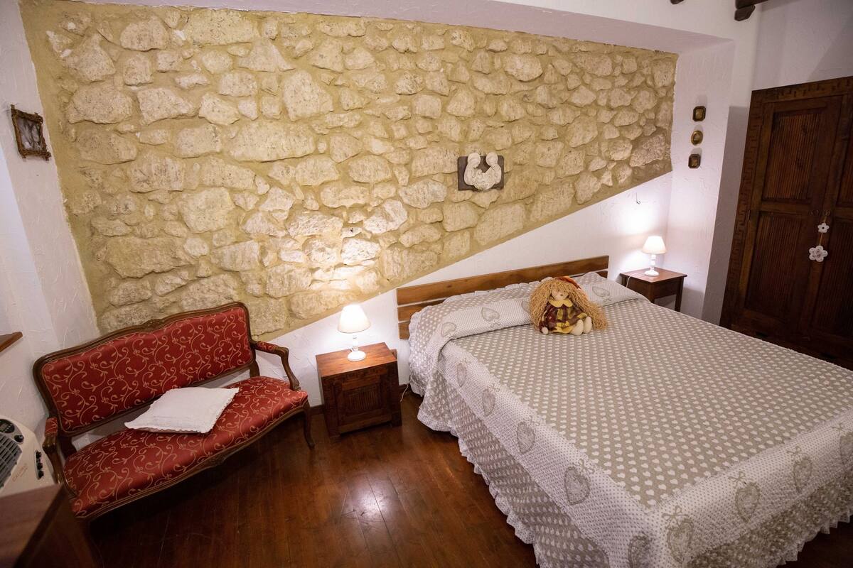 Un dormitorio con pared de piedra, una cama de madera, un sofá rojo y una mesita de noche con una lámpara.