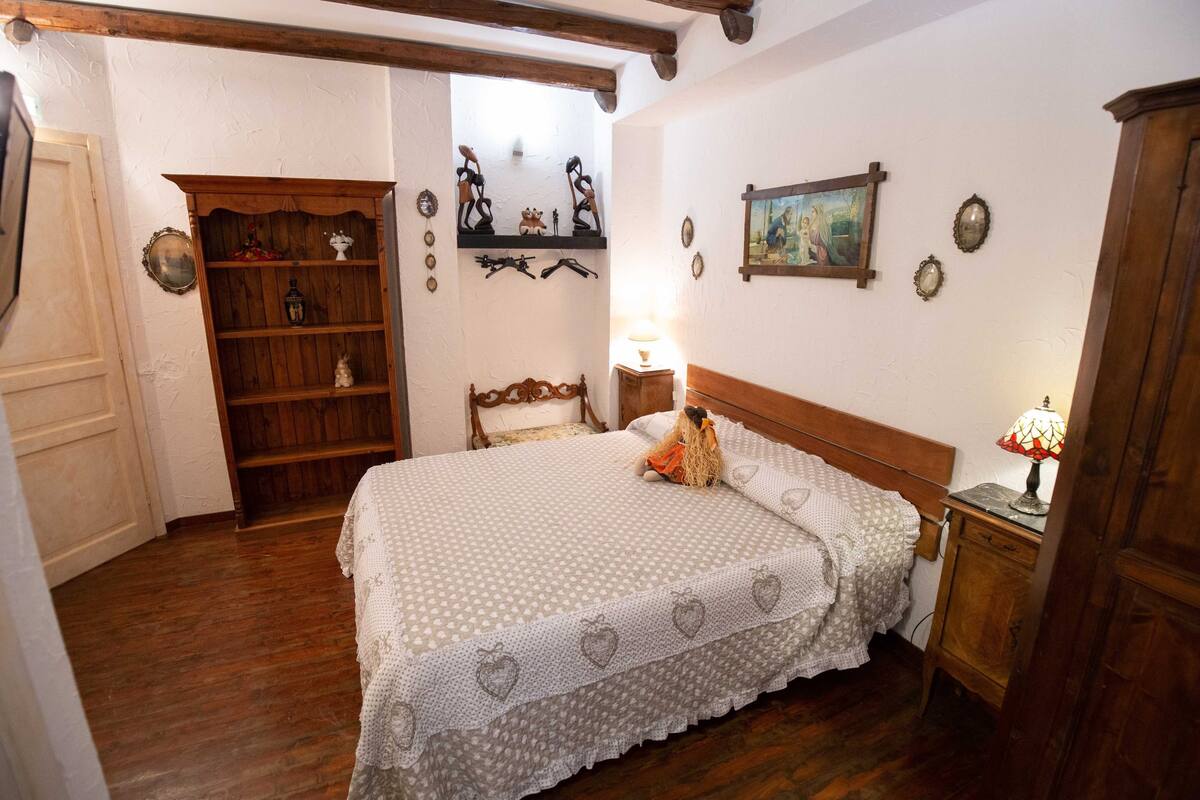 Un dormitorio con una cama de madera, una mesita de noche con lámpara, un armario de madera y una estantería con objetos decorativos.
