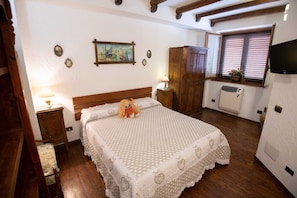 A bedroom with a wooden bed, a nightstand with a lamp, a dresser, a window with blinds, and a television mounted on the wall.