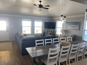 A modern living room with a blue sectional sofa, a wooden dining table with white chairs, and a ceiling fan.
