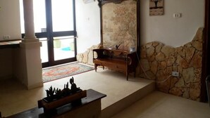 Una habitación con pared de piedra, un banco de madera, una pequeña mesa con un tablero de ajedrez y un ventanal con vistas.