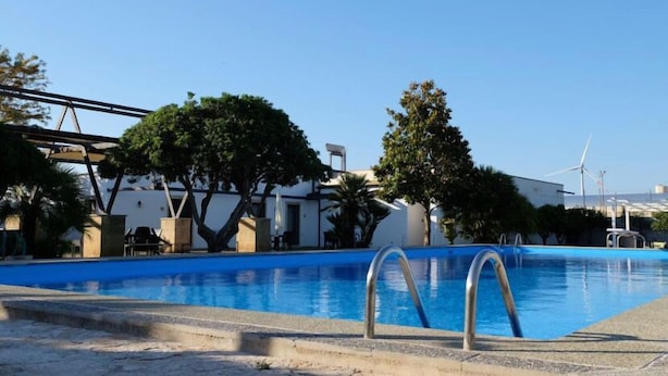 Una piscina con agua cristalina azul, rodeada de árboles y un edificio de paredes blancas y techo negro.