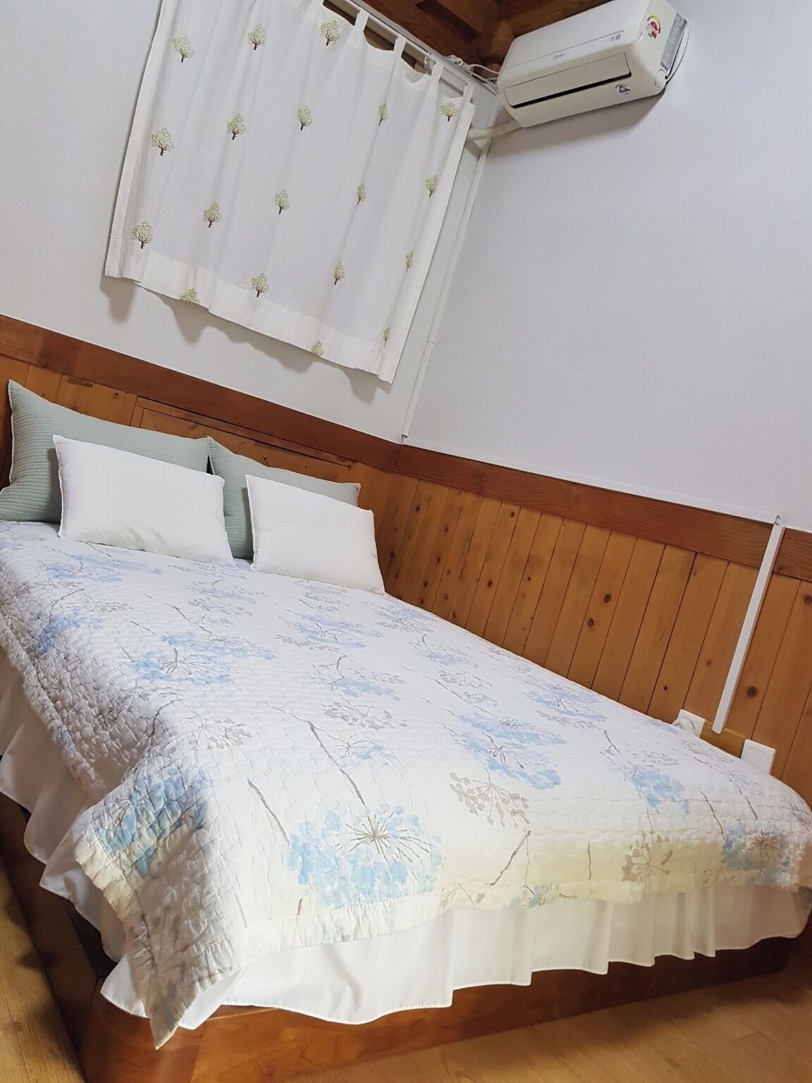 Un dormitorio con cabecera de madera, cama con colcha estampada con flores, almohadas blancas y cortina blanca con diseño de árbol.