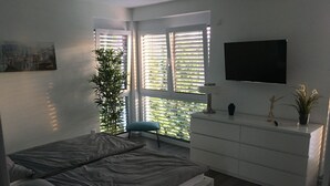 A modern bedroom with a bed, a dresser, a TV, and a window with blinds.