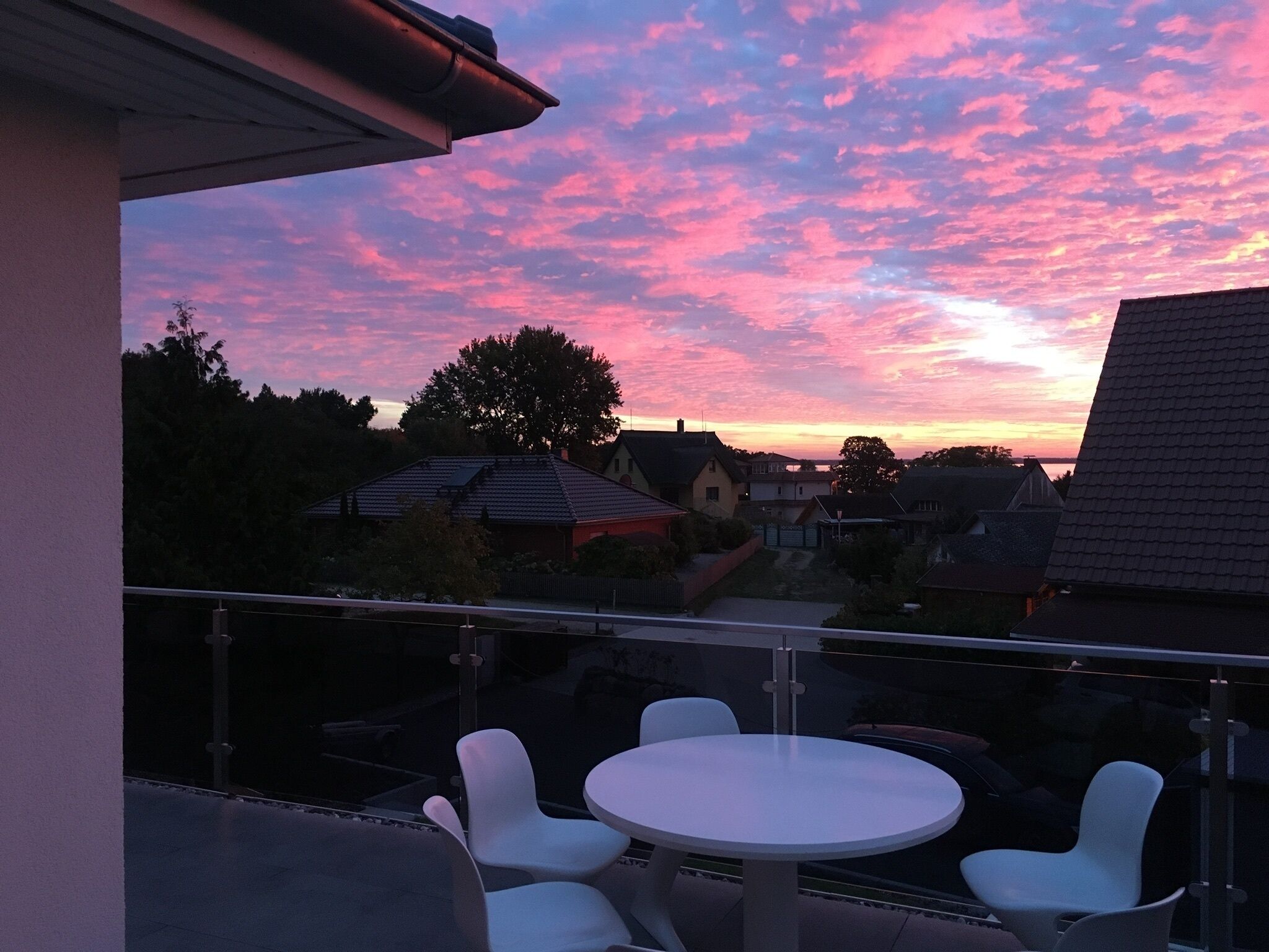 A balcony with a table and chairs, overlooking a residential area with houses and trees during sunset.