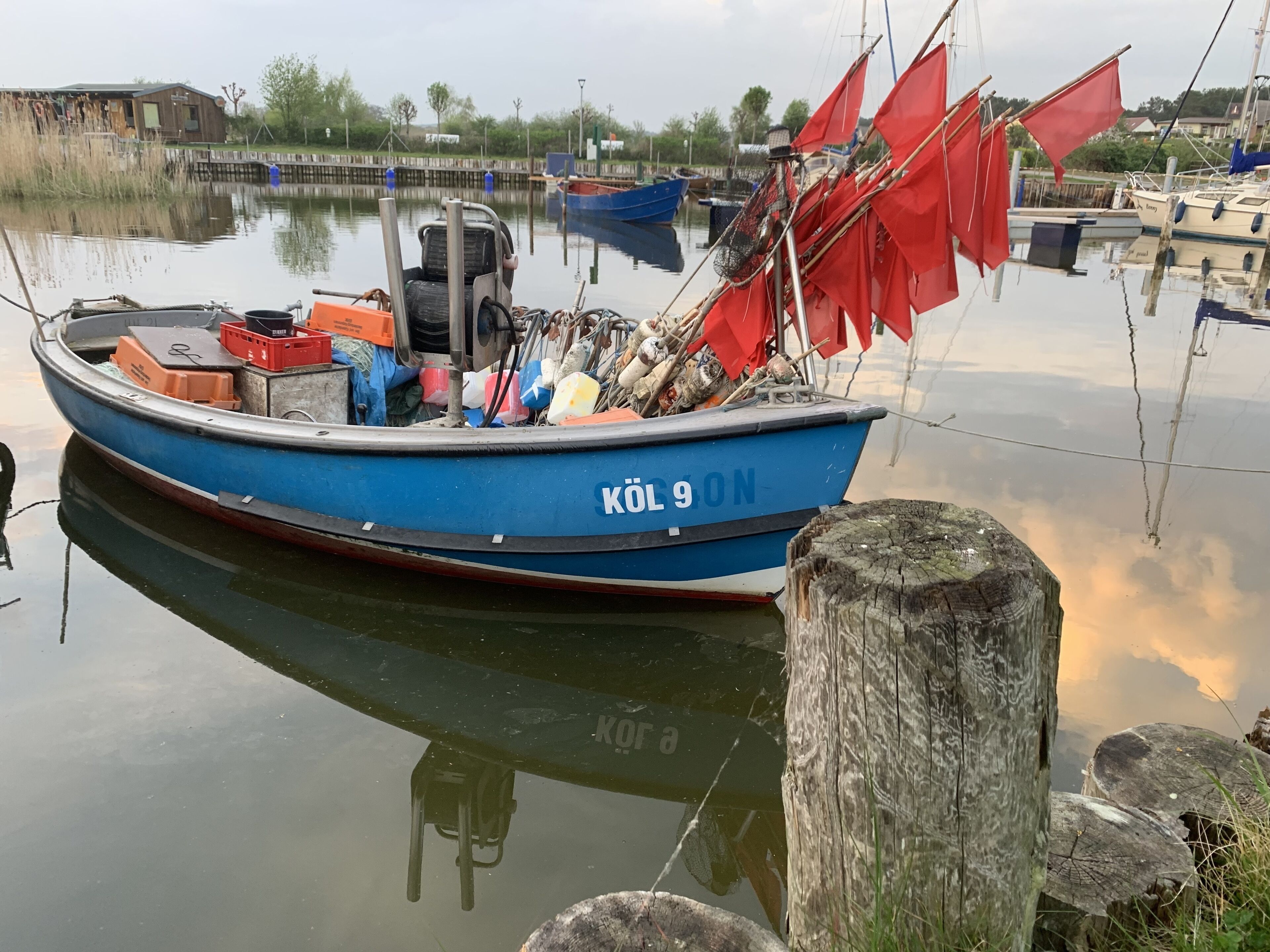 A blue boat with the number "KÖL 9" docked at a calm water body, surrounded by wooden posts and other boats.
