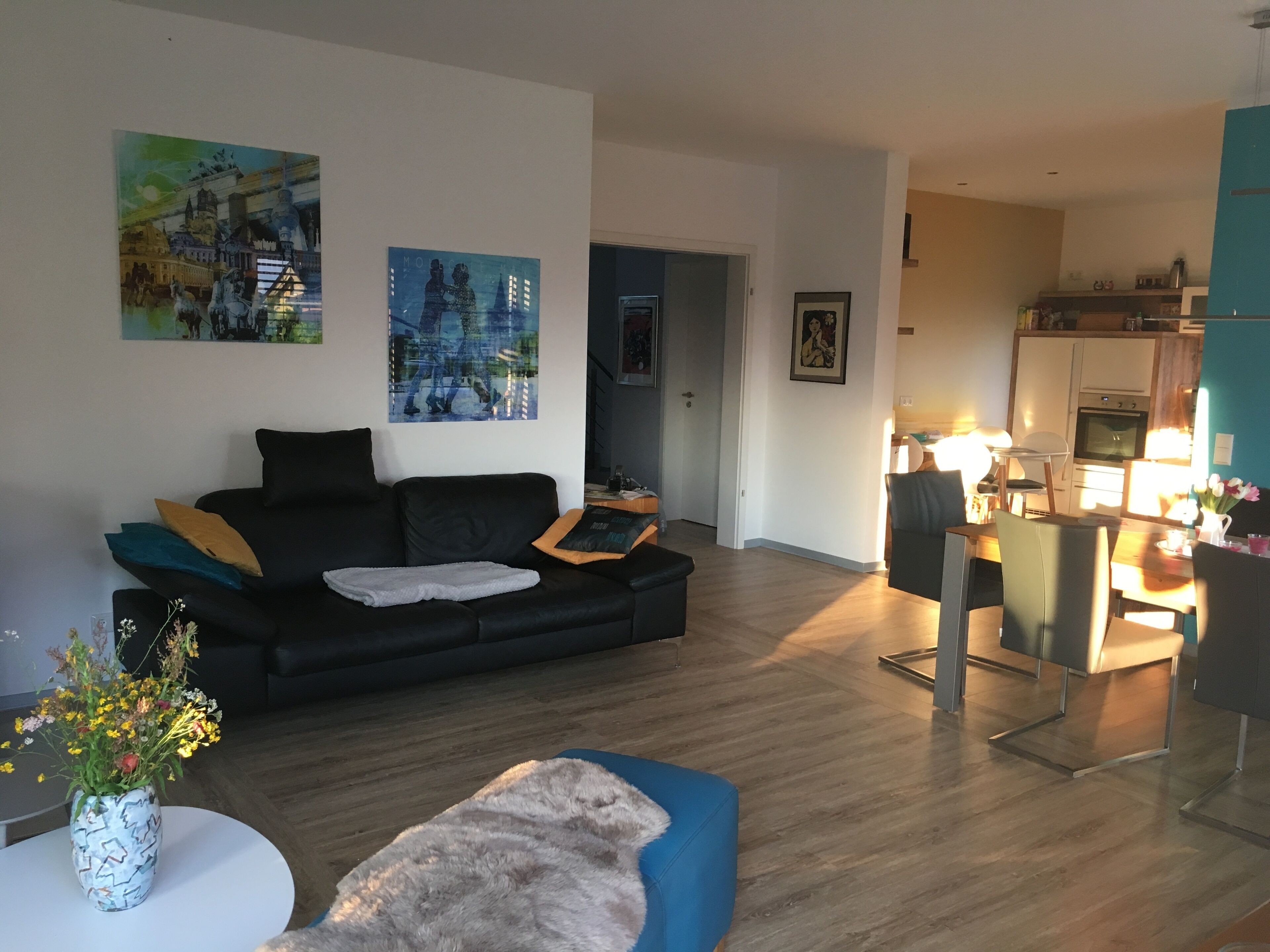 Un salon moderne avec un canapé noir, un pouf bleu et une table ronde blanche ornée d’un vase de fleurs. Deux tableaux sont accrochés au mur, il y a un coin cuisine avec une table et des chaises, ainsi qu’un couloir qui mène à une autre pièce.