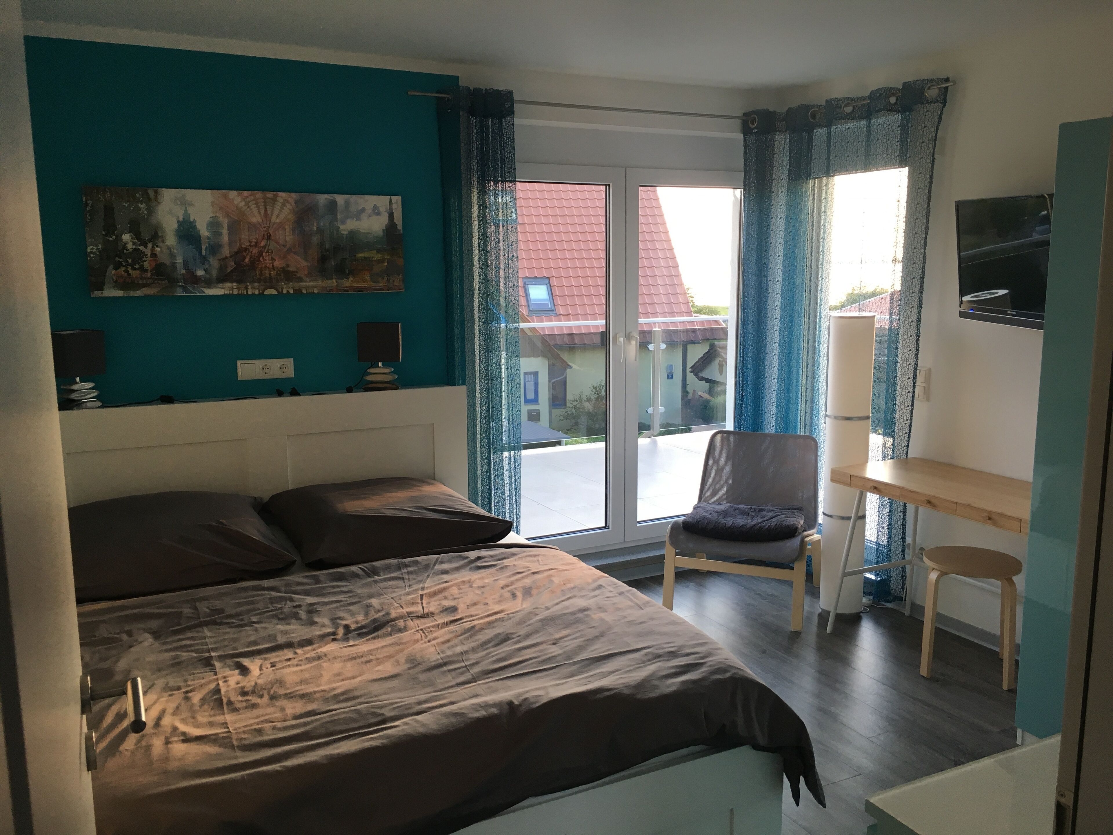 Een slaapkamer met een bed, een bureau, een stoel, een schilderij en uitzicht op een huis door het raam.