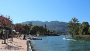 A riverside walkway with boats docked, a jet ski, and mountains in the background. - Lungolago by Interhome (Lavena Ponte Tresa)