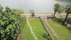 A garden with a stone pathway leading to a seating area by a lake, surrounded by greenery and palm trees. - Lungolago by Interhome (Lavena Ponte Tresa)