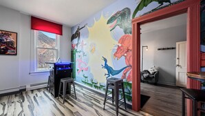 Une pièce décorée d’une fresque représentant des dinosaures, une fenêtre avec un store rouge et un coin bar avec des tabourets.