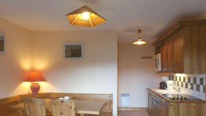 Une cuisine chaleureuse, avec des meubles en bois, une table à manger et un banc. Il y a une lampe sur la gauche et une cuisinière sur la droite.