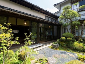 Ein traditionelles japanisches Gebäude mit modernem Glaseingang, umgeben von einem gepflegten Garten mit Steinen, Moos und verschiedenen Pflanzen.