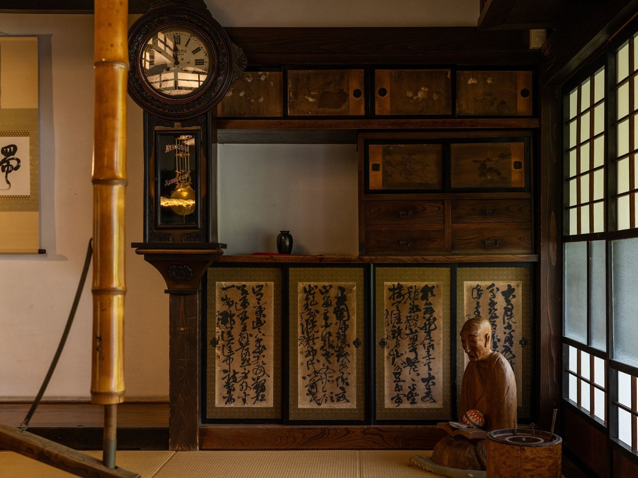 Une pièce de style japonais traditionnel, avec un sol recouvert de tatamis, une horloge en bois, des œuvres de calligraphie et une statue.