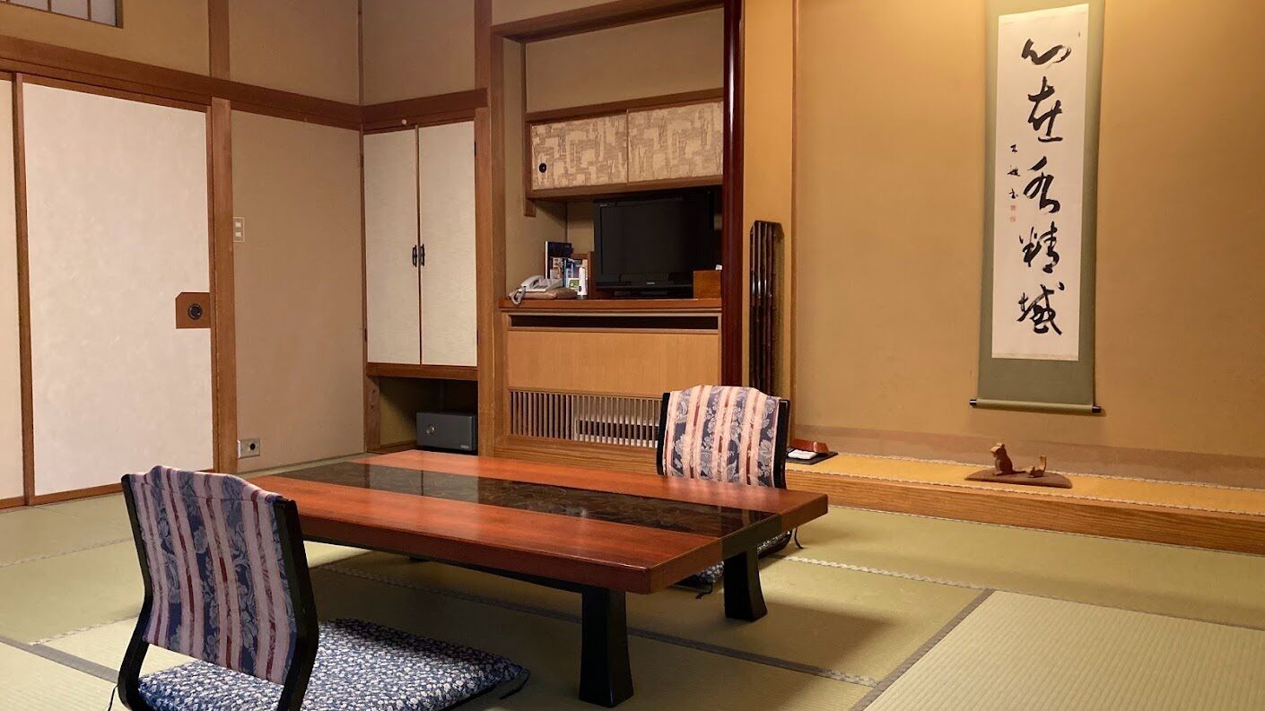 Um quarto japonês tradicional com tatames, uma mesa baixa, duas cadeiras estampadas, uma televisão e um pergaminho de caligrafia na parede.
