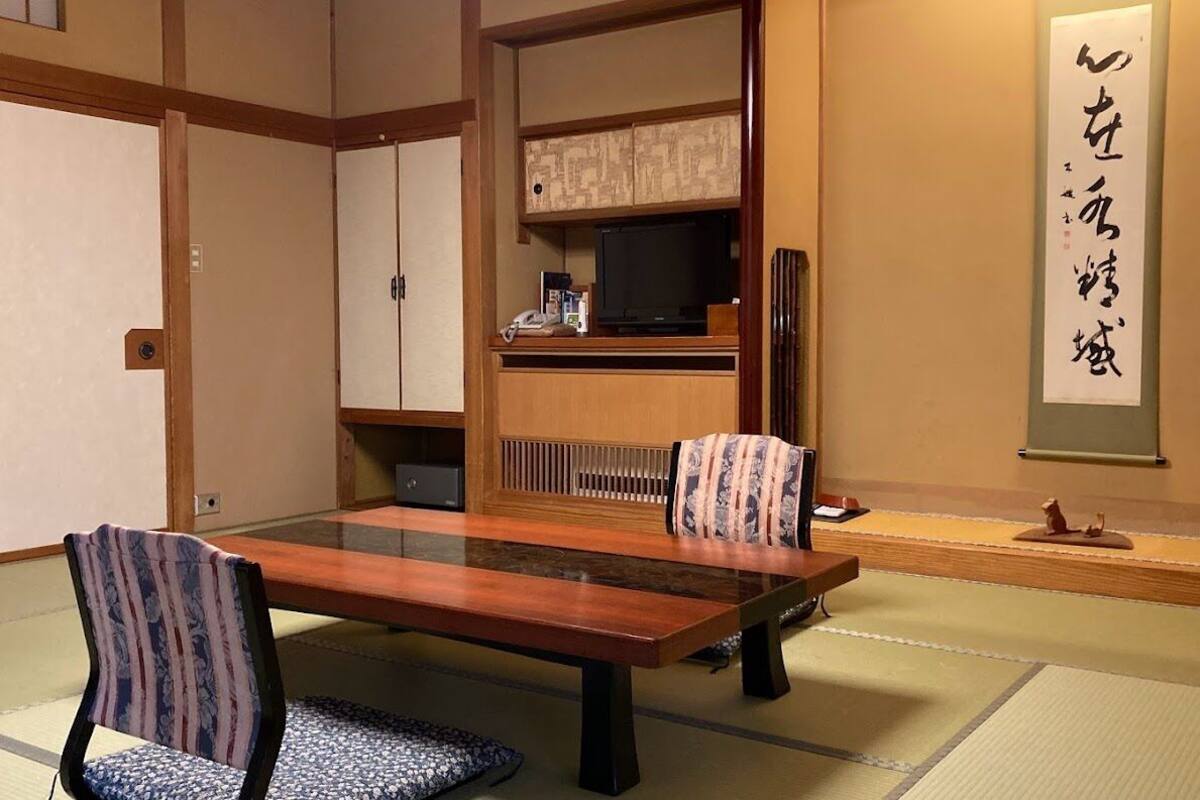 A traditional Japanese room with tatami flooring, a low table, two patterned chairs, a television, and a calligraphy scroll on the wall.