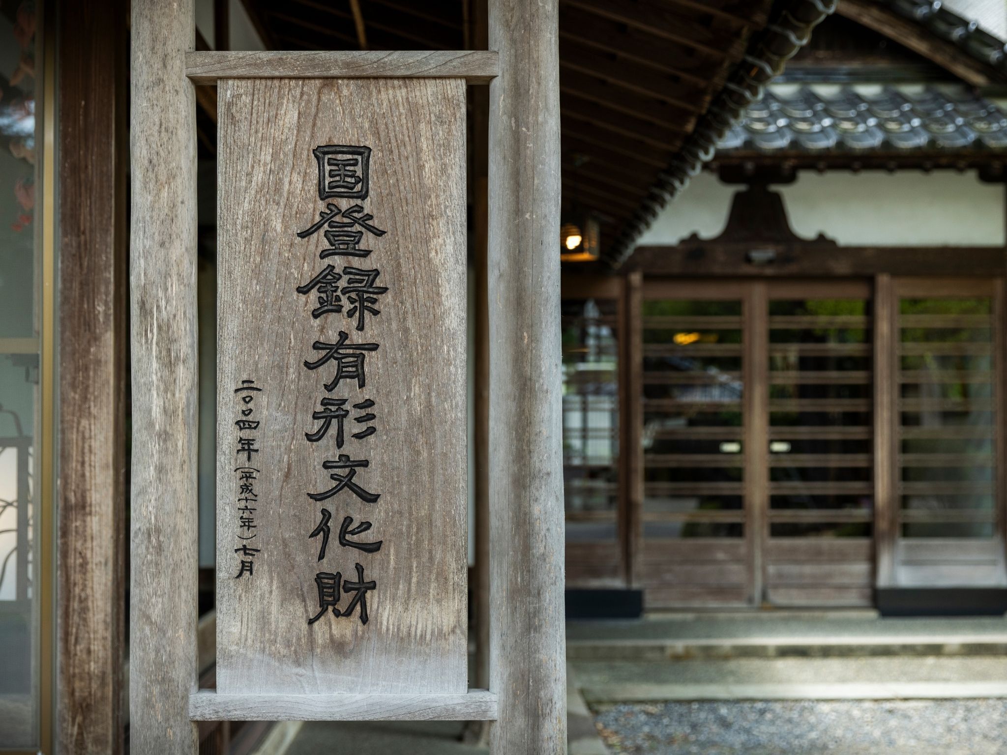 Un letrero de madera con caracteres japoneses, parte de una construcción tradicional con tejado de azulejos y puertas de madera.