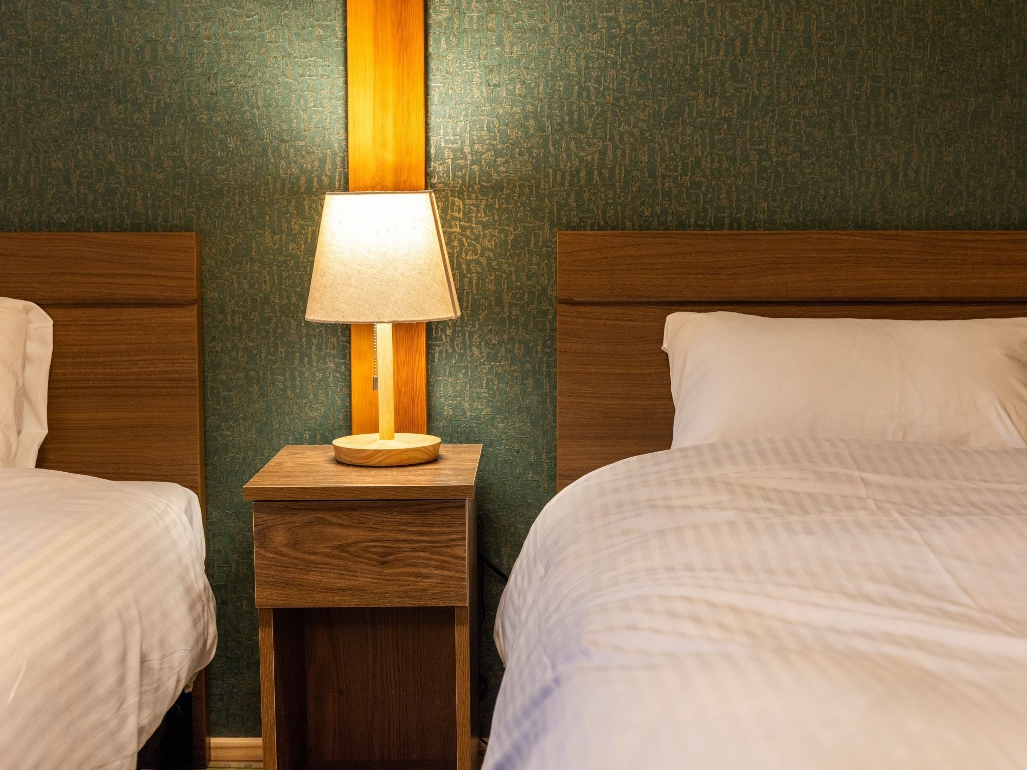 A hotel room with two beds, a nightstand with a lamp, and a green textured wall.