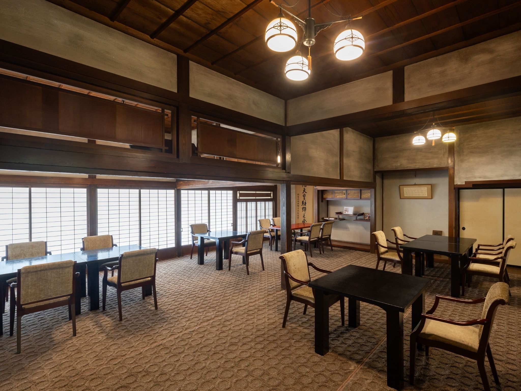 A spacious room with multiple tables and chairs, large windows, and a high ceiling with hanging lights.