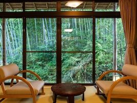 Quarto com uma janela ampla que oferece vista para uma floresta de bambu. Há duas cadeiras de madeira e uma mesa redonda pequena.