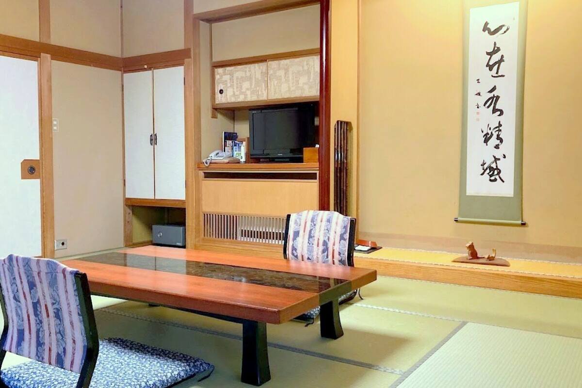 A traditional Japanese room with tatami flooring, a low wooden table, two chairs, a television, and a calligraphy scroll on the wall.