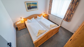 A double bed with white linens, two bedside tables with lamps, a painting on the wall, and a window with curtains. - Haus Strandruh (Binz)