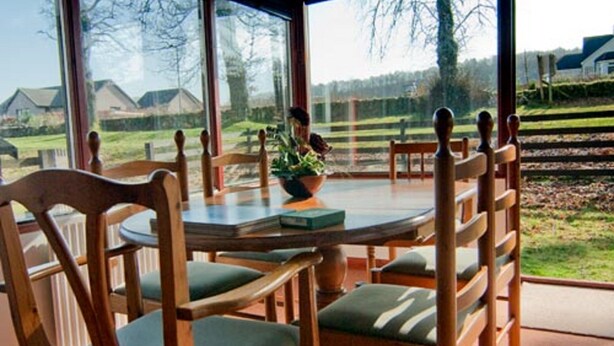 Een eethoek met een ronde tafel, houten stoelen met groene kussens en uitzicht op een woonwijk door grote ramen.
