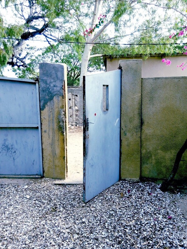 A metal gate with a blue door, set in a gravel area with a stone wall and greenery in the background. - Auberge Cœur Océan (Joal-Fadiout)