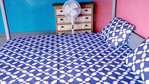 Ein Bett mit kariertem blau-weißem Bettbezug, eine Holzkommode mit mehreren Schubladen und ein Ventilator in der Ecke.
